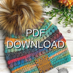 May include: A colorful, hand-crocheted beanie with a large, fluffy faux fur pom-pom. The hat features a mix of vibrant colors and textures, with the words "PDF DOWNLOAD" prominently displayed. A small leather tag reads "SWEATER WEATHER".