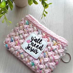 May include: A pink crocheted drink sleeve with a white patch that reads "y'all need Jesus" in black script. The sleeve has a textured, bubble-like pattern with colorful flecks and a silver key ring.