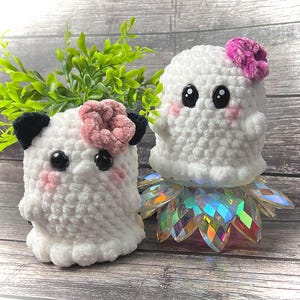 May include: Two white crocheted ghost plushies with black eyes and pink cheeks. One ghost has a pink flower on its head and the other has a black and white flower on its head.