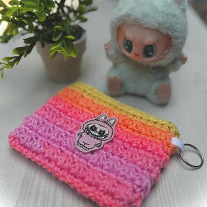 May include: A handmade crochet wallet in a gradient of yellow, orange, and pink. A small patch of a cartoon bunny is on the front. A small plush doll in a bunny costume is in the background.