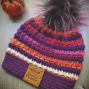 May include: A crocheted beanie featuring horizontal stripes in purple, orange, pink, and cream. The hat is topped with a dark purple and pink faux fur pom-pom and includes a square tan leather tag with the text Fall is my second favorite F-word.