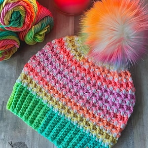 Könnte beinhalten: Eine bunte gehäkelte Mütze mit einem Regenbogenstreifenmuster und einem flauschigen Bommel. Die Mütze ist aus verschiedenen leuchtenden Farben gefertigt, darunter Pink, Orange, Gelb, Grün und Blau. Der Bommel ist eine Mischung aus Weiß, Pink und Orange. Die Mütze liegt auf einer Holzoberfläche.
