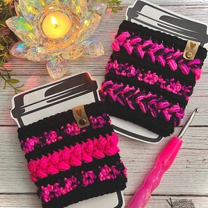 May include: Two black and pink crocheted coffee cup cozies with a heart-shaped tag. The cozies are on a white wooden surface with a pink crochet hook and a candle in a clear glass lotus holder.