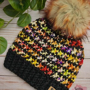 Könnte beinhalten: Handgehäkelte Mütze mit schwarzem Rand und farbenfrohem, strukturiertem Oberteil. Das Oberteil weist eine Mischung aus roter, gelber, orangefarbener und violetter Wolle auf. Ein großer, flauschiger Kunstfell-Bommel in Braun- und Cremetönen bedeckt die Mütze. Ein kleines Etikett mit Text ist angebracht.