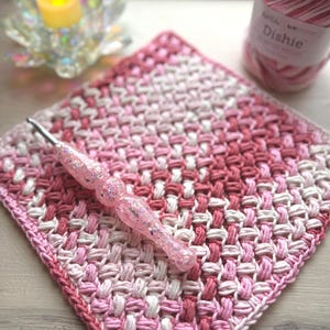 May include: A pink and white crocheted square with a matching crochet hook. A ball of pink and white yarn is in the background. The crochet hook has a glittery handle. The square has a textured pattern and a pink border.