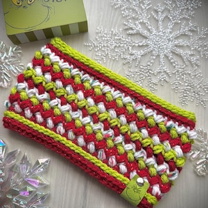 May include: A woven headband in red, white, and green. The headband has a lime green border and a small green tag with a face on it. A box with the Grinch's face and the words "STINK, STANK, STUNK" is in the background.