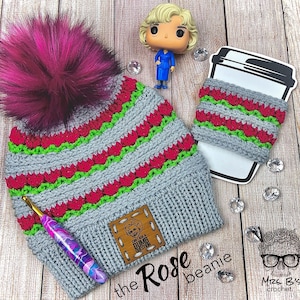 May include: A gray and pink crocheted beanie with a pom pom on top. The beanie has a green and pink striped pattern and a wooden tag that says "Can't I get a Dumb Question?" The beanie is on a wooden surface with a crocheted coffee cup cozy next to it.