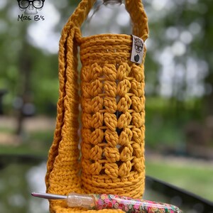 May include: A yellow crocheted bottle holder with a long strap. The bottle holder is made with a stitch pattern that resembles a flower. The holder is being used to hold a clear plastic water bottle. A crochet hook is also pictured.