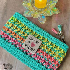 May include: A teal crochet pouch with a rainbow stripe pattern. The pouch has a zipper closure and a small pink and teal tassel charm. The pouch has a white patch with the text "Take care of yourself" and a red heart.