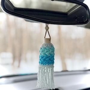 May include: A handmade car air freshener with a gradient blue and white crochet design. The bottom features a white fringe, and it hangs from a wooden bead and string. The air freshener is suspended in front of a car's rearview mirror.