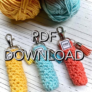 Könnte beinhalten: Drei gehäkelte Lippenbalsam-Halter in Gelb, Blau und Koralle mit passenden Quastenanhängern. Die Halter sind an silbernen Schlüsselringen befestigt. Der Text "PDF DOWNLOAD" befindet sich in der Mitte des Bildes.
