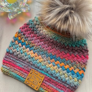 May include: A multi-coloured knitted beanie hat with a large faux fur pom-pom. The hat has a woven pattern in shades of blue, pink, orange, and green. A small, brown leather tag with the text "Yes, I'm cold" is sewn on the side. The hat is designed for cold weather.
