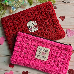 May include: Two crocheted zippered pouches, one red and one pink. The red pouch has a Snoopy character patch and the pink pouch has a patch that says "Take care of yourself" with a heart. Both pouches are on a wooden surface with hearts scattered around them.