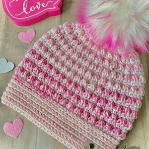 May include: Pink and white crocheted beanie with a fluffy white and pink pom pom. The beanie has a pink and white striped band at the bottom. The beanie has a small tag that says "Mrs. B's".