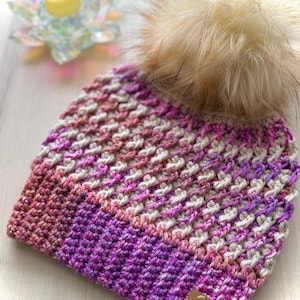 May include: A hand-knitted beanie in shades of purple, pink, and white, with a fluffy, light brown pom-pom. The hat has a textured, woven pattern and a small leather tag. The hat is a winter accessory.