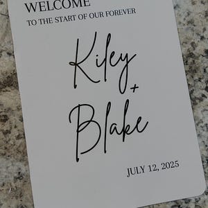May include: A white wedding fan with black text. The fan reads "WELCOME TO THE START OF OUR FOREVER" above the names "Kiley + Blake" and the date "JULY 12, 2025". The fan has a wooden handle.
