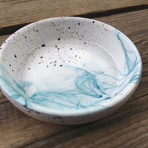 May include: A small white ceramic bowl with a blue and black marbled design. The bowl has a speckled white glaze with black dots and blue swirls.