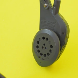 K&ouml;nnte beinhalten: Ein dunkelgraues elektronisches Kopfh&ouml;rer-Ohrst&uuml;ck mit einem kreisf&ouml;rmigen Lautsprechergitter mit mehreren Perforationen, einem seitlichen Lautst&auml;rkeregler und eingepr&auml;gtem Text, der besagt, dass das Ger&auml;t Teil 15 der FCC-Regeln entspricht.