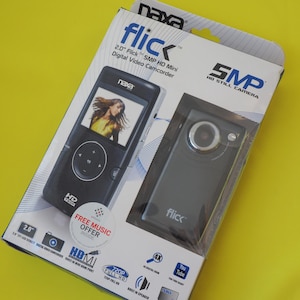 May include: A Naxa Flick 2.0-inch 5MP HD mini digital video camcorder in its original retail packaging. The box features product imagery, the Naxa logo, and text including 5MP HD Still Camera, 720P Full HD, and a Free Music Offer sticker.