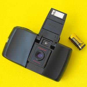 May include: A black Kodak film camera with a pop-up flash and an Ektanar 35mm f/4.5 lens, positioned next to a yellow and black CR123A battery. The camera body features the Kodak brand name embossed on the flash unit and red text on the lens.