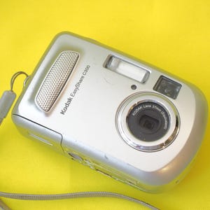 May include: A silver Kodak EasyShare C300 digital camera with a textured silver shutter button, a black lens housing labeled Kodak Lens 37mm Equivalent, a flash unit, and a light gray wrist strap attached to the side of the camera body.