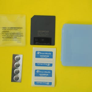 Könnte beinhalten: Eine Sammlung von SmartMedia-Zubehör, darunter eine schwarze Olympus-Speicherkarte, eine blaue Kunststoffhülle, eine transparente Kunststoffhülle mit Text, ein Streifen silberner Kreise und ein Bogen SmartMedia-Etiketten mit blauem und weißem Text, alles auf gelbem Hintergrund.