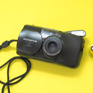 May include: A black Olympus Stylus Zoom 200 Panorama film camera with a 35-70mm lens, a black wrist strap, and a single CR123A lithium battery positioned to the right of the camera body. The camera features a sliding lens cover and a built-in flash.