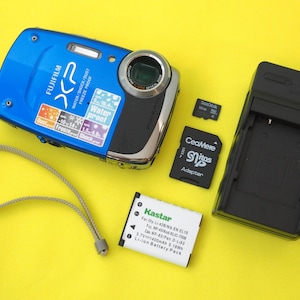 May include: A bright blue Fujifilm XP digital camera with a wrist strap, a black battery charger, a white Kastar lithium-ion battery pack, a black CeaMere SD adapter, and a small SanDisk memory card. The camera features waterproof, shockproof, and dustproof labels.