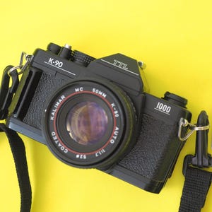 May include: A black Kalimar K-90 1000 TTL 35mm film camera with a 50mm 1:1.7 lens, featuring a black textured body and a black neck strap. Next to the camera is a small blister pack containing two round silver LR44 button cell batteries.