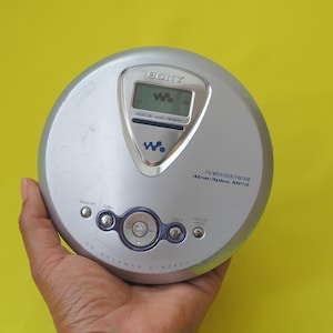 Puede incluir: Reproductor de CD Sony plateado con pantalla digital. El dispositivo tiene botones para funciones de radio y memoria. El texto "TV/WEATHER/FM/AM Atrac3plus MP3" está impreso en el dispositivo. El Walkman se sostiene en una mano sobre un fondo amarillo.