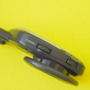 K&ouml;nnte beinhalten: Ein dunkelgraues tragbares Radio mit seitlichem Abstimmrad und einem Bandschalter mit der Beschriftung FM, DX, LOCAL und AM. Das Ger&auml;t verf&uuml;gt &uuml;ber ein schwarzes Kabel und einen runden Lautsprecher, wobei das Wort TUNING in Wei&szlig; aufgedruckt ist.