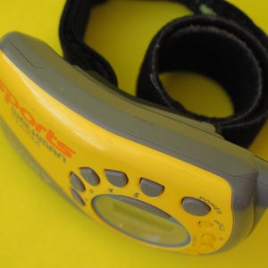 Puede incluir: Un Sports Walkman amarillo y gris con una correa negra. El dispositivo tiene varios botones y una peque&ntilde;a pantalla. El texto "Sports Walkman" est&aacute; impreso en naranja en la parte delantera. Sobre un fondo amarillo.