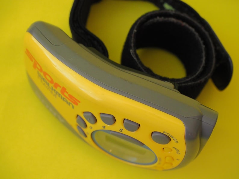 Puede incluir: Un Sports Walkman amarillo y gris con una correa negra. El dispositivo tiene varios botones y una peque&ntilde;a pantalla. El texto "Sports Walkman" est&aacute; impreso en naranja en la parte delantera. Sobre un fondo amarillo.