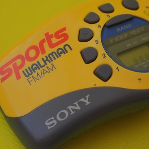 Puede incluir: Radio Sony Sports Walkman FM/AM amarilla y gris. Tiene una pantalla digital y botones de preajuste. La palabra "Sports" es roja, y "Walkman" es azul.