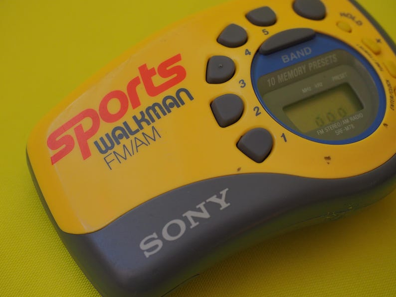 Puede incluir: Radio Sony Sports Walkman FM/AM amarilla y gris. Tiene una pantalla digital y botones de preajuste. La palabra "Sports" es roja, y "Walkman" es azul.