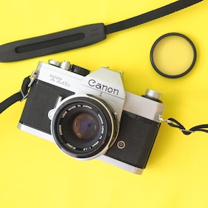 May include: A silver and black Canon TLb camera with a lens, strap, and filter on a yellow background. The camera lens is labeled "Canon Lens Made in Japan". The camera strap is black.