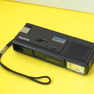 May include: A black Keystone XR308 Telephoto Everflash camera with a rectangular body, a built-in flash unit on the right side, a black wrist strap, and white and blue text printed on the top surface indicating the model and brand name.