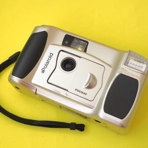 May include: A silver Polaroid PDC640 digital camera with black rubberized grip accents on the sides. The front features a central lens, a flash unit on the right, and a black wrist strap attached to the left side of the camera body.