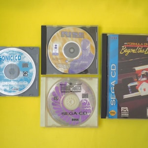 Puede incluir: Cuatro cajas de videojuegos SEGA CD sobre un fondo amarillo. Los juegos incluyen "Sonic CD", "Rise of the Robots", "Double Switch" y "Formula One Beyond the Limit". Los discos del juego son visibles dentro de las cajas.