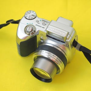 May include: A silver Olympus SP-510UZ digital camera featuring a black grip, a prominent lens, and a pop-up flash unit. The camera includes a black neck strap attached to the sides and displays the brand name Olympus on the front housing.