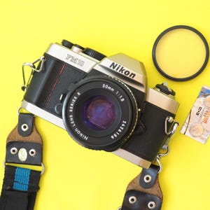 May include: A silver and black Nikon FM10 35mm film camera with a 50mm 1:1.8 lens, a black camera strap with blue accents, a circular lens filter, and a small package of two Maxell button cell batteries on a bright yellow background.