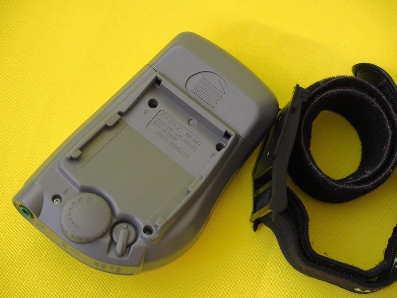 Puede incluir: Radio Sony SRF-M33 gris con correa negra, sobre fondo amarillo. La radio tiene un compartimento para pilas y un dial de control. La correa es de material negro suave.