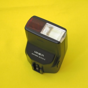Puede incluir: Un flash de cámara Minolta Maxxum 2000i negro con un cuerpo rectangular, un tubo de flash de vidrio transparente a la derecha y una ventana de sensor de color rojo oscuro a la izquierda. El nombre de la marca y el modelo están impresos en blanco.