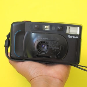 May include: A black Fuji point-and-shoot film camera featuring a Fujinon lens, a built-in flash, and a red shutter button on the top right. The camera includes a black wrist strap attached to the side and displays the Fuji logo on the front.