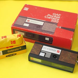 May include: A vintage GAF Pocket Camera 220 in its original red box, with a brown and black camera and a roll of Kodak Kodacolor II film. The camera has a black strap and is on a yellow background.