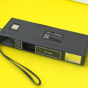 May include: A black Vivitar 700 film camera with a rectangular body, a silver shutter button, a built-in flash, and a black wrist strap. The front features the Vivitar brand name, 24mm f/5.6 lens specifications, and a small window for the viewfinder.