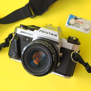 Könnte beinhalten: Eine silberne und schwarze Pentax Super Program 35mm Filmkamera mit einem 50mm Objektiv, schwarzem Gurt und einer Packung Maxell LR44 Batterien auf gelbem Hintergrund. Das Kameraobjektiv ist mit SMC Pentax-A 1:1.7 beschriftet.
