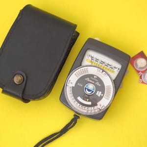 May include: A Gossen Luna Pro light meter with a circular dial, a needle display, and a black wrist strap, accompanied by a black leather carrying case with a snap button closure and two small round silver batteries on red backing cards.