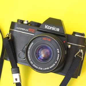 May include: A black Konica Autoreflex TC camera with a 40mm lens and a black strap. The lens is marked "KONICA HEXANON AR 40mm F1.8 LENS MADE IN JAPAN 55ø". The camera is set against a yellow background.