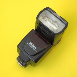 Peut inclure: Un flash d'appareil photo Nikon Speedlight SB-700 noir doté d'une tête inclinable rectangulaire avec un panneau avant transparent, une fenêtre d'affichage teintée de rouge sur le corps principal et une griffe de fixation métallique.